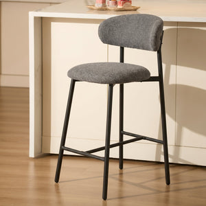 A modern bar stool with a gray upholstered seat and curved backrest, supported by a black metal frame, set in a bright kitchen environment.
