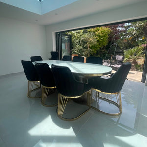 A modern dining room featuring a round white marble-top dining table with six black upholstered chairs that have gold metal wire bases, set in a bright room with a large skylight and glass doors overlooking a garden.