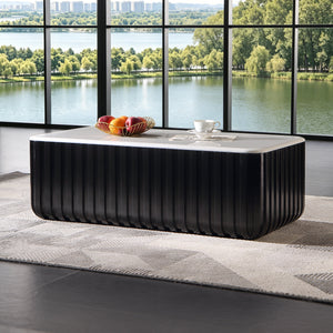 A modern black ribbed rectangular coffee table with a white top sits on a gray patterned rug in front of a large black-framed window overlooking a lake and green trees.