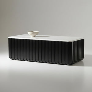 A modern black ribbed rectangular coffee table with a white top sits on a gray patterned rug in front of a large black-framed window overlooking a lake and green trees.