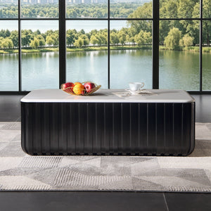 A modern black ribbed rectangular coffee table with a light gray stone top sits in a room with a large floor-to-ceiling window overlooking a lake and green trees. A fruit bowl and a white teacup are placed on the table.