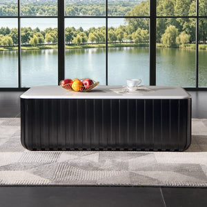 A modern black ribbed rectangular coffee table with a light gray stone top sits in a room with a large floor-to-ceiling window overlooking a lake and green trees. A fruit bowl and a white teacup are placed on the table.