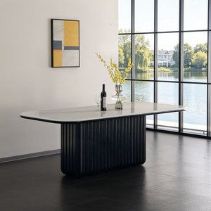 A modern dining table with a ribbed black base and a light gray stone top, styled in a minimalist room with a large window overlooking water.