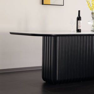 A modern black ribbed pedestal dining table with a white curved tabletop, styled in a minimalist room with a wine bottle, wine glass, and vase on top.