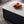 A modern rectangular coffee table with a glossy white top and a dark charcoal fluted base, styled with a gold wire fruit bowl on a gray patterned rug.