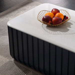 A modern rectangular coffee table with a glossy white top and a dark charcoal fluted base, styled with a gold wire fruit bowl on a gray patterned rug.