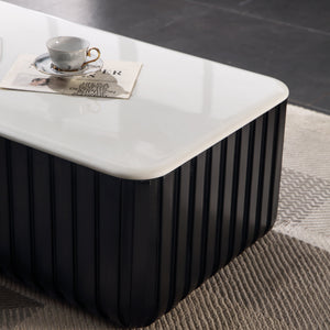 A modern rectangular coffee table with a glossy white top and a black ribbed pedestal base, styled on a textured beige rug with a coffee cup on top.