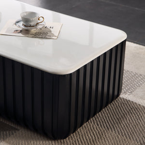 A modern rectangular coffee table with a glossy white top and a black ribbed pedestal base, styled on a textured beige rug with a coffee cup on top.