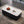 A modern rectangular coffee table with a white top and a dark ribbed base, styled on a geometric rug in a contemporary room with large windows overlooking a lake and forest.