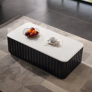 A modern rectangular coffee table with a white top and a dark ribbed base, styled on a geometric rug in a contemporary room with large windows overlooking a lake and forest.