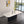 A modern rectangular dining table with a glossy white top and a fluted black pedestal base, styled in a minimalist room with a large window overlooking water.