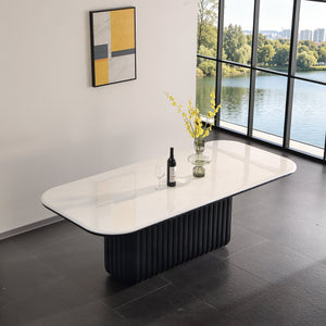 A modern rectangular dining table with a glossy white top and a fluted black pedestal base, styled in a minimalist room with a large window overlooking water.