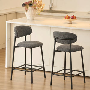Two modern bar stools with textured gray fabric seats and backs on black metal frames, positioned in front of a white kitchen island with a sink and countertop.