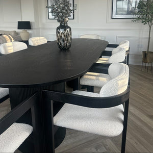 A modern dining room setup featuring a large oval black table with textured white boucle chairs, styled with a dark patterned vase of dried foliage in the center.