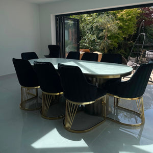 An oval dining table with a white marble-look top and gold metal base, surrounded by eight black velvet chairs with gold wire bases, set in a modern dining room with large glass doors opening to a garden.