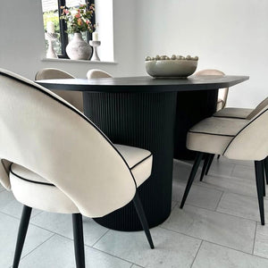 A modern dining room setup featuring a round black ribbed pedestal table surrounded by cream upholstered dining chairs with black legs and piping, set on light gray tiled flooring.