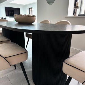 A modern round dining table with a dark wood top and a ribbed cylindrical pedestal base in black, surrounded by upholstered dining chairs in a bright dining room.