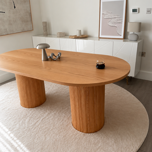 An oval wooden dining table with fluted pedestal legs, styled in a modern room on a round cream rug.