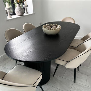 A modern dark wood oval dining table with a fluted pedestal base, surrounded by six cream upholstered dining chairs with black legs, set in a contemporary dining room with light gray tiled flooring.