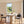 A modern dining room and kitchen interior with a round white pedestal table surrounded by gray upholstered chairs, plus a wooden island with three bar stools. Large windows reveal greenery outside, and the room has warm wood flooring and beige walls.
