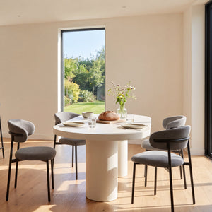 A modern dining room and kitchen interior with a round white pedestal table surrounded by gray upholstered chairs, plus a wooden island with three bar stools. Large windows reveal greenery outside, and the room has warm wood flooring and beige walls.
