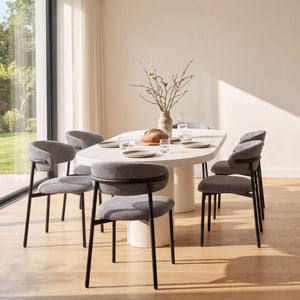A modern dining room scene with a round white dining table, six gray upholstered chairs with black metal legs, and a matching kitchen island with three gray bar stools. Large windows reveal a green garden outside, and the room has light wood flooring and warm beige walls.