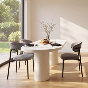 A modern dining room scene with a round white dining table, four gray upholstered chairs with black legs, and a kitchen island with four matching bar stools. Large windows reveal greenery outside, and the room is lit by warm natural sunlight.