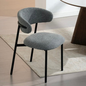 A modern gray upholstered dining chair with a curved backrest and black metal legs, shown in a bright interior setting next to a round table.