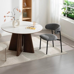 A modern round dining table with a white marble-look top and dark wood pedestal base, styled in a bright room with two upholstered chairs on a light rug.