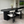 A modern dining room setup featuring a dark oval dining table with a ribbed cylindrical pedestal base, surrounded by four white upholstered chairs with black frames. The room has light beige walls, a large skylight, and a built-in entertainment unit in the background.