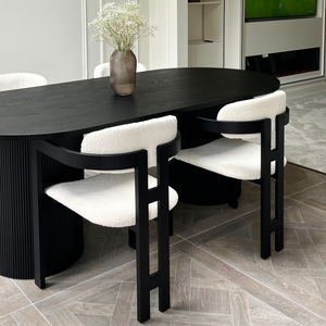 A modern dining table set in a contemporary dining room. The set features a dark oval-shaped table with a fluted base and four matching chairs upholstered in a textured cream bouclé fabric.