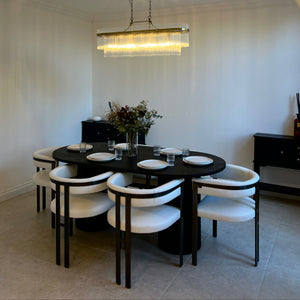 A modern dining room setup featuring a round black dining table with six white upholstered chairs, a rectangular glass chandelier, and a warm beige arched ceiling.