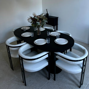 A modern dining room setup featuring a black oval table surrounded by six white upholstered chairs with black metal frames, set under a rectangular glass chandelier.