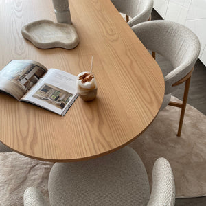 A modern dining setup featuring a light oak wood oval table surrounded by four textured cream boucle chairs with light wood legs. An open magazine and a small drink with a straw rest on the table, set on a beige rug over dark wood flooring.