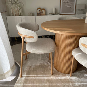 A modern dining set featuring a round light wood table and three upholstered chairs with textured cream fabric and light wood legs, set on a neutral rug in a contemporary dining room.