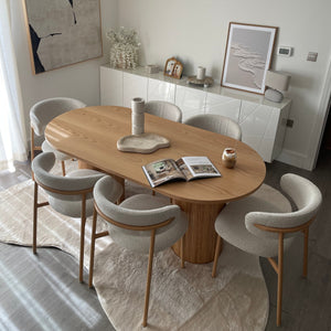 A modern dining room setup featuring a round light oak wood table surrounded by six white upholstered chairs with curved backs and light wood legs, set on a textured white rug.