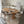 A modern dining room setup featuring a round light oak wood table with six matching upholstered chairs in a light gray fabric, set on a large round textured cream rug. The room has light gray wood floors, white walls, and minimalist framed wall art.