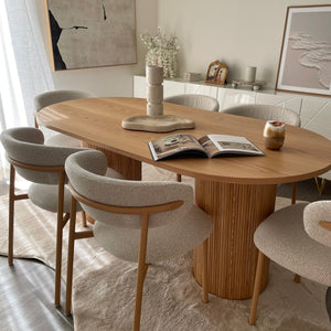 A round wooden dining table with a slatted pedestal base, surrounded by upholstered dining chairs in a bright, modern dining room setting.
