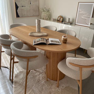 A modern dining room setup featuring a round light oak wood table with a slatted pedestal base, surrounded by upholstered dining chairs in a neutral beige and white color palette, set on a textured white rug.