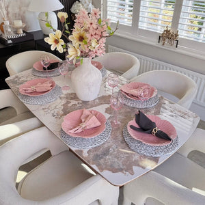 Dining table set with pink plates and napkins, white vase with flowers, and white chairs in a bright room.