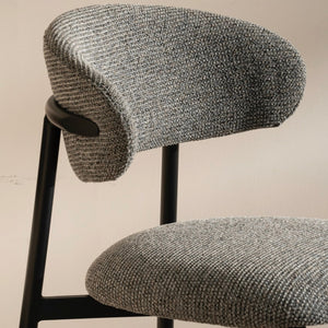 A close-up of a modern bar stool featuring a textured gray woven fabric seat and curved backrest, supported by a slim black metal frame against a plain beige wall.