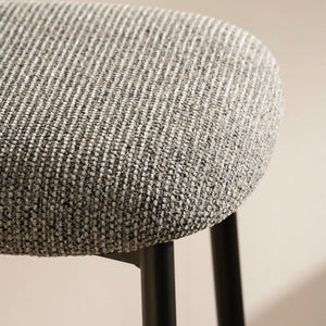 Close-up of a modern stool with a textured gray and white woven fabric seat cushion and slim black metal legs, set against a warm beige background.