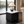A modern black pleated side table with a white marble top, styled in a contemporary living room with a lit table lamp on top, set against a large window overlooking water.