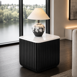 A modern black pleated side table with a white marble top, styled in a contemporary living room with a lit table lamp on top, set against a large window overlooking water.