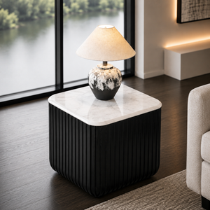 A modern square side table with a black pleated base and a white marble top, styled in a contemporary living room with a table lamp on top.