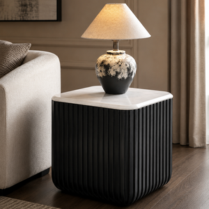 A modern black side table with vertical fluting and a white marble top, styled in a living room with a lit table lamp on top.