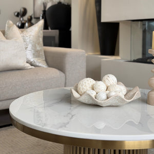 A modern living room scene featuring a round marble-top coffee table with a gold base, holding a decorative bowl of white seashells. In the background, a light gray fabric sofa with textured pillows is visible next to a fireplace and framed wall art.