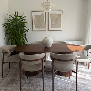 A modern dining room setup featuring a dark wood oval dining table surrounded by six upholstered dining chairs with dark wood frames, set on a distressed gray area rug. A white ceramic vase sits on the table, and a large green potted plant is on the left.