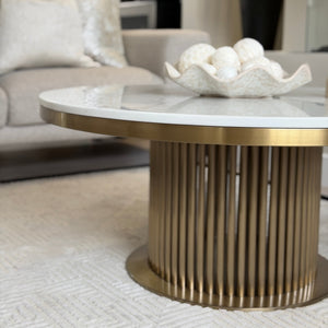 A modern round coffee table with a white marble top and a fluted gold metal base, styled in a contemporary living room setting.