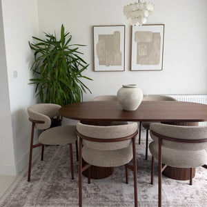 A dining room scene showing a round wooden dining table with upholstered chairs, a white ceramic vase on top, a green potted plant, framed wall art, and a patterned rug against a white wall.